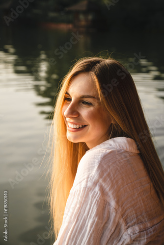 Young smiling woman sitting by the lake at sunset, summer evening portrait on vacation. © sav_an_dreas