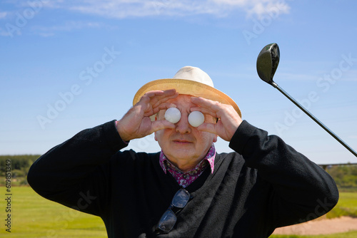 Senior man holds golf balls in front of his eyes against the background of a golf course and golf club. Concept of golf crazy , crazy about golf or golf mad. photo