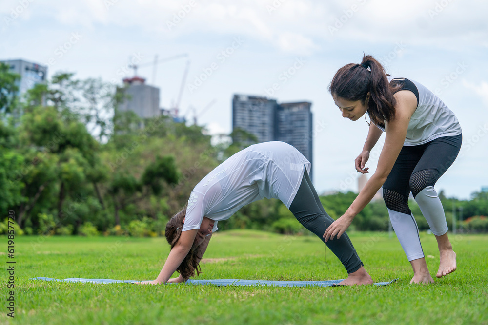 Young Indian female yoga teacher wearing sportswear trains eight-year-old girl to yoga poses on a mat to stretch her legs in the park.