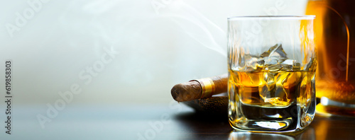 A bottle and a glass of whiskey with ice and a steaming Cuban cigar on a table against a light background. Men's club banner idea. Copy space for text photo