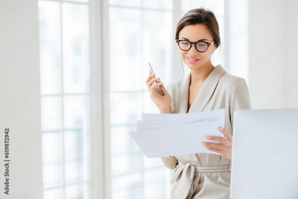 Satisfied woman in business suit, working in office, holding cellphone, wearing glasses, pleased expression Satisfied woman in business suit, working in office, holding cellphone, wearing glasses, pleased expression