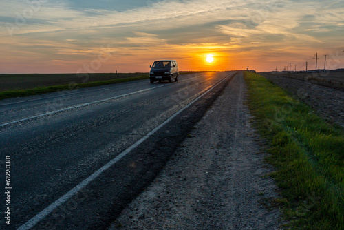 The silhouette of a car on the road against the background of the sun. The car is driving on the highway during sunset. The concept of travel and freedom. © Pokoman