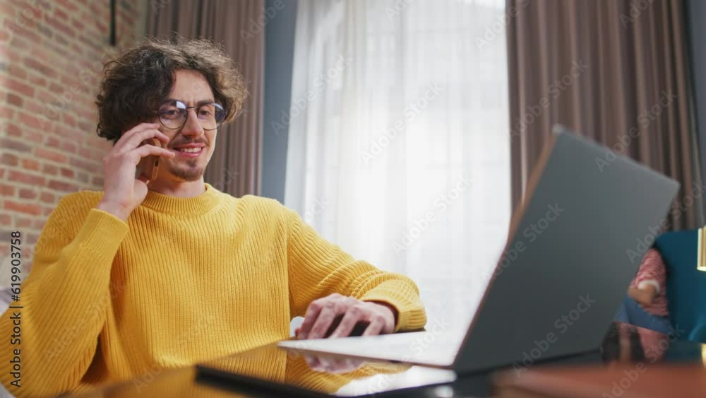 Caucasian man speaking on his phone while using laptop in room. In background his loving wife having videocall on tablet device. People enjoying remote communication with their friends or relatives. Caucasian man speaking on his phone while using laptop in room. In background his loving wife having videocall on tablet device. People enjoying remote communication with their friends or relatives.
