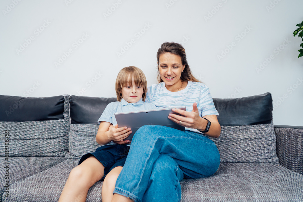Mother and son on the sofa using digital tablet. Happy mom and little boy using touch screen tablet for watching videos, reading books, playing video game, using online app, studying together at home