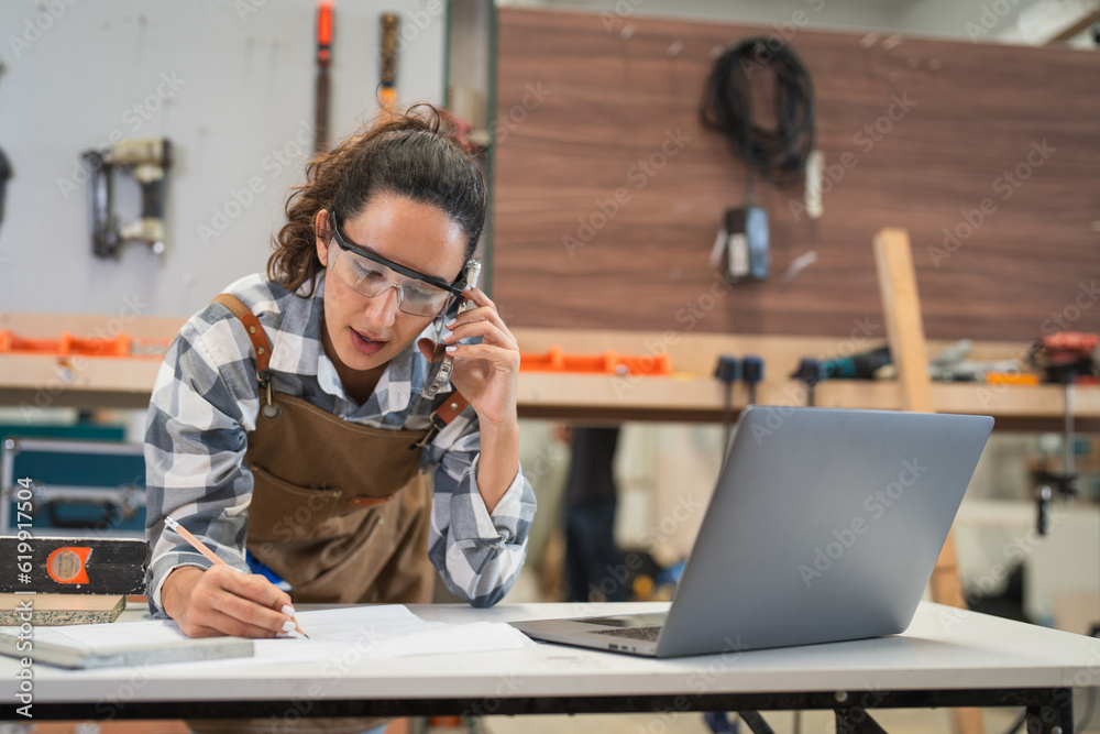 Female carpenter using laptop computer and talking with client by smartphone at carpentry workshop. Woman wearing glasses safety working with wood at furniture factory - obrazy, fototapety, plakaty 