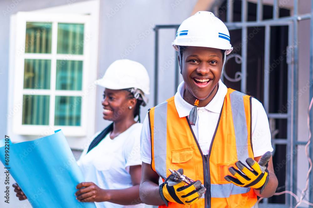 Male and female workers in hard hats standing in front of a building smiling Male and female workers in hard hats standing in front of a building smiling