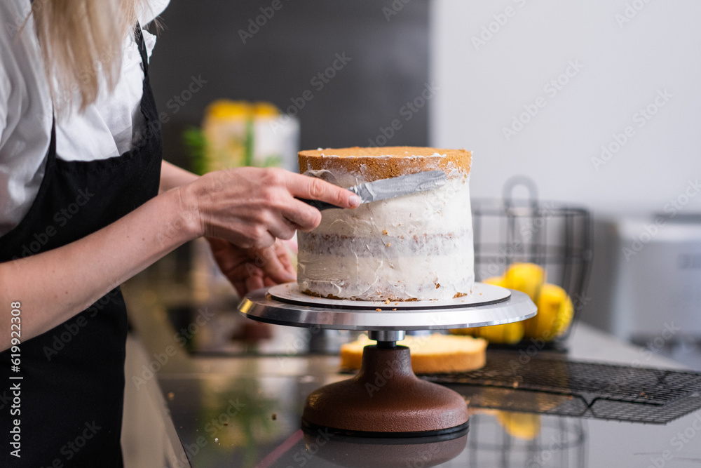 Experienced self-made confectioner smearing tasty cream around baked cake layers with knife woman in black apron showing detailed process of making delicious pastry on confectionery courses