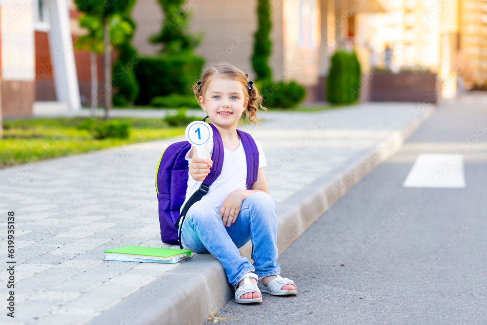 a schoolgirl child with a backpack and bills in her hands sits on the curb at the school and shows the number one, the concept is back to school