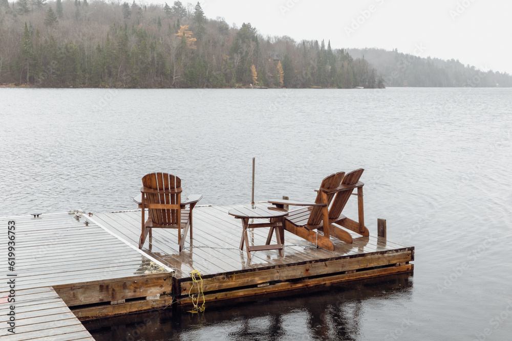 Obraz premium trois chaises en bois sur un quai sur l'eau au bord d'un lac avec une forêt de l'autre côté lors d'une journée brumeuse