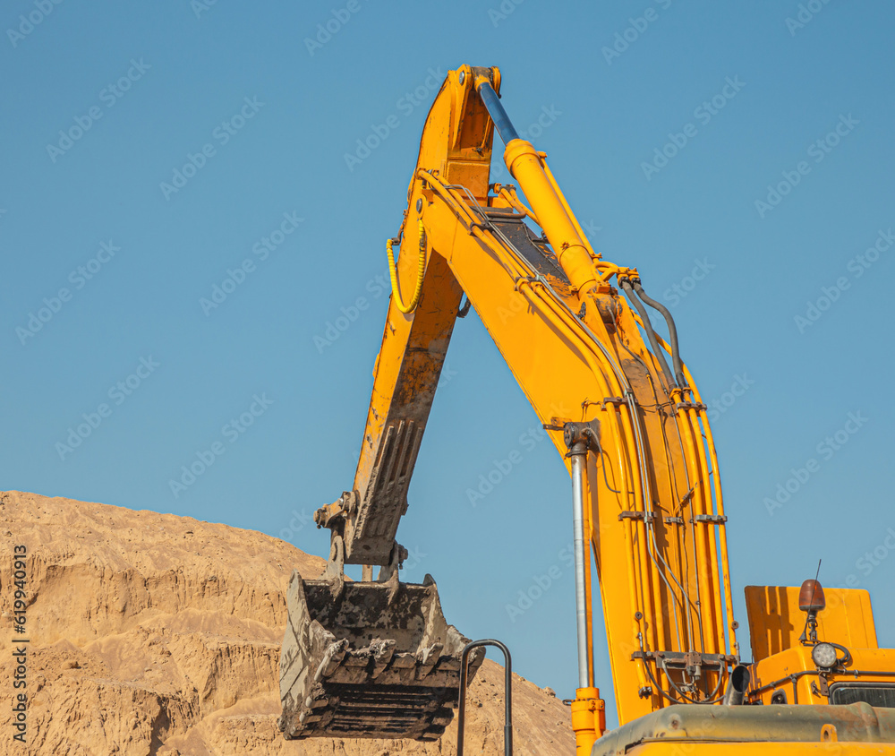 Excavator with bucket while working against the background of blue sky and yellow sand pile close-up. Excavator bucket on the background of a pile of sand in the quarry. Excavator with bucket while working against the background of blue sky and yellow sand pile close-up. Excavator bucket on the background of a pile of sand in the quarry.