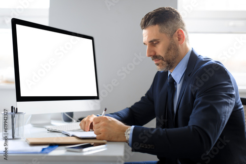 Serious middle aged businessman at table with computer with empty screen making notes, working in office, mockup