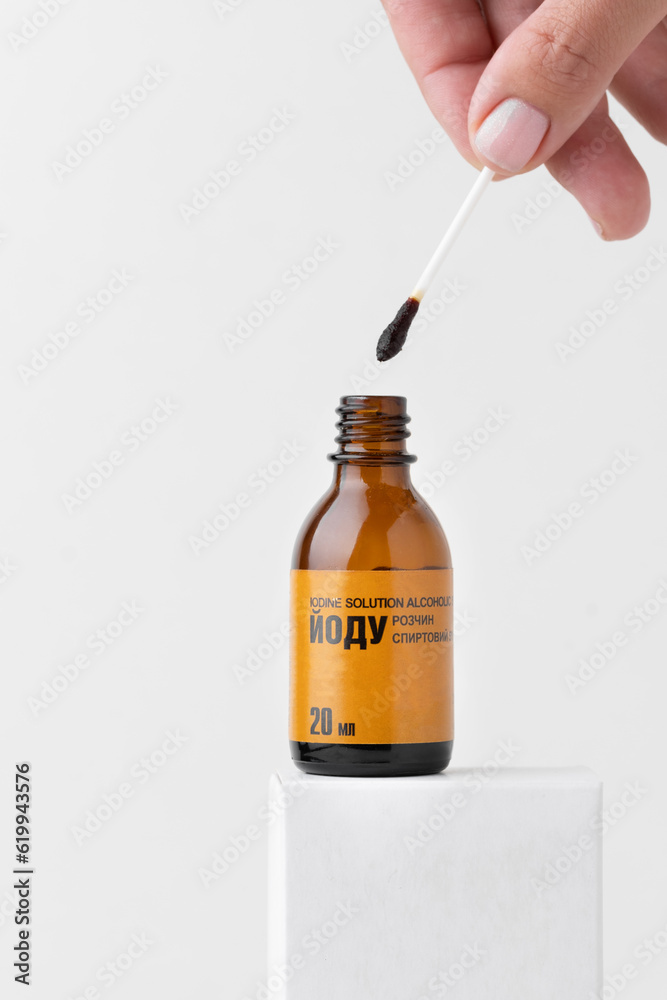 Woman dipping cotton bud into glass bottle with medical iodine on the white background. Close up of brown glass bottle with alcohol solution of iodine inscription in Ukrainian. Woman dipping cotton bud into glass bottle with medical iodine on the white background. Close up of brown glass bottle with alcohol solution of iodine inscription in Ukrainian.