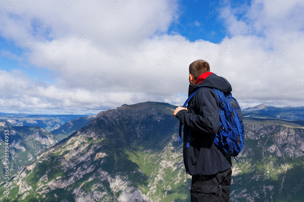 An unrecognisable man in a black jacket with a blue backpack stands on a cliff and looks at the mountains. The concept of an active lifestyle
