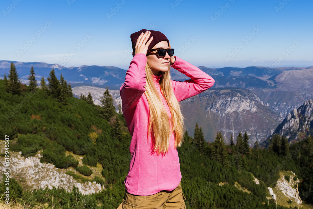 Portrait of a young woman mountain guide in glasses, hat and pink jacket on the background of mountain peaks in sunny weather