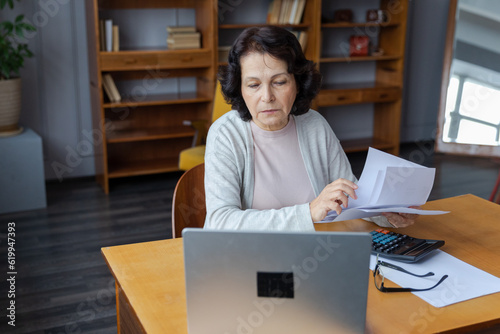 Middle aged senior woman sit with laptop and paper document, Pensive older mature lady reading paper bill pay online at home managing bank finances calculating taxes planning loan debt pension payment