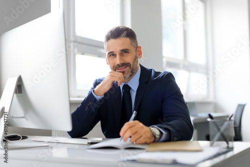 Portrait of happy middle aged man entrepreneur working on computer and taking notes, sitting at workplace and smiling