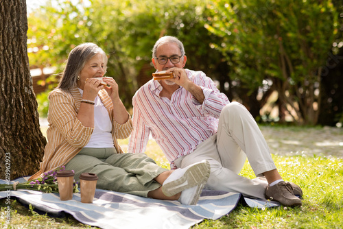 Positive old european husband and wife eat sandwich, enjoy picnic together in park, lunch outdoor