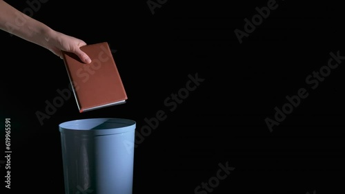 Bin with outdated book. A view of person hand throwing the book into the plastic bin on the black background.