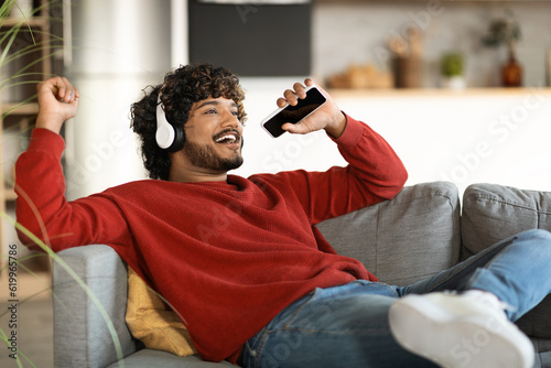 Positive Indian Guy Singing While Relaxing With Smartphone And Headphones At Home