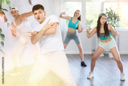 Positive smiling male teenager learns social dance, perform movements in choreographic class in company of peers. Young girls in ball cap and guy repeat movements, train in spacious training hall.