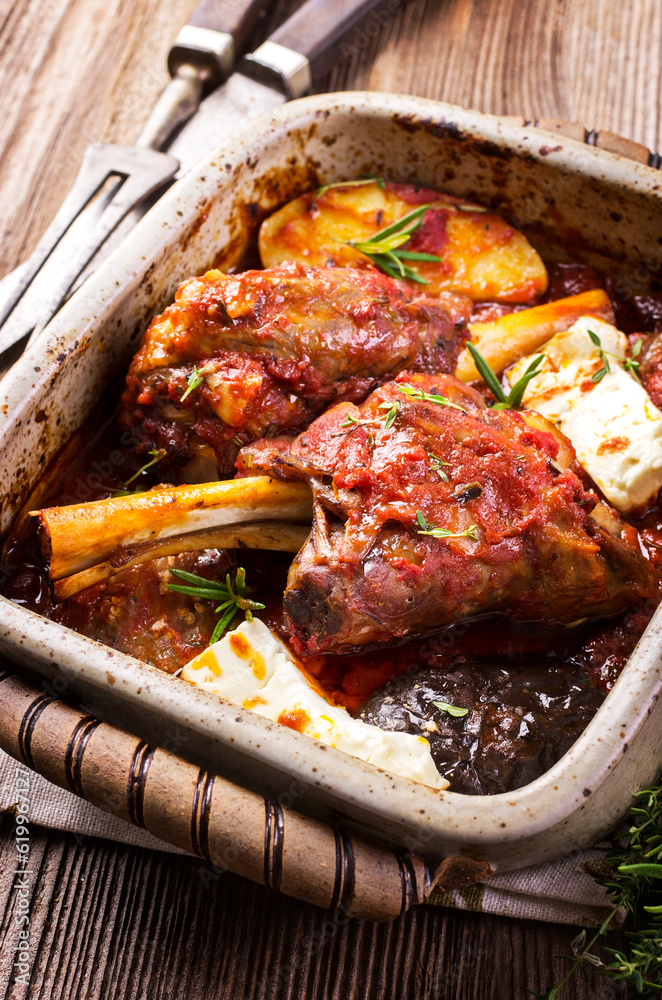 Traditional Greek braised lamb knuckle with feta cheese and eggplant in spicy tomato sauce served as close-up on a rustic roasting pan