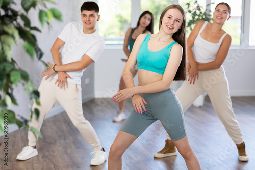 Smiling teenage girl dancer practicing active vigorous dance with group of friends in modern studio..