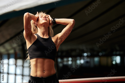 Young athlete girl getting ready for a workout at the stadium. Attractive slender blonde in a black tracksuit straightens and pins up her long hair before training. Active lifestyle and sports.