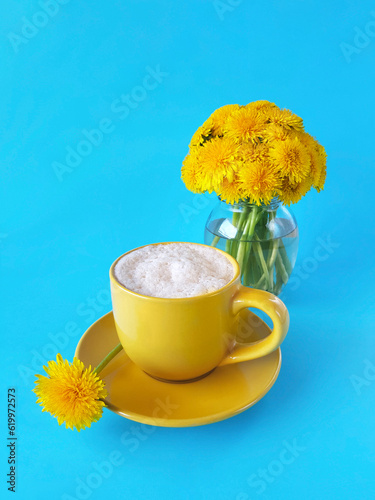 Coffee topped with whipped cream in yellow cup and one dandelion on saucer. Bouquet of fresh dandelions (Taraxacum officinale) in little glass vase on blue backgroung. Hello summer concept. Vertical.