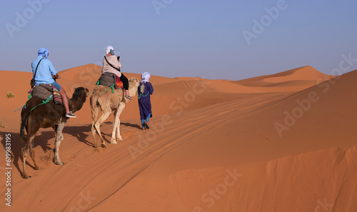 Tourists ride camels through the sand dunes in Sahara Desert. Berber man in national clothes leading camel caravan with tourists near village Merzouga  Morocco. Taken at sunset with orange - red sands