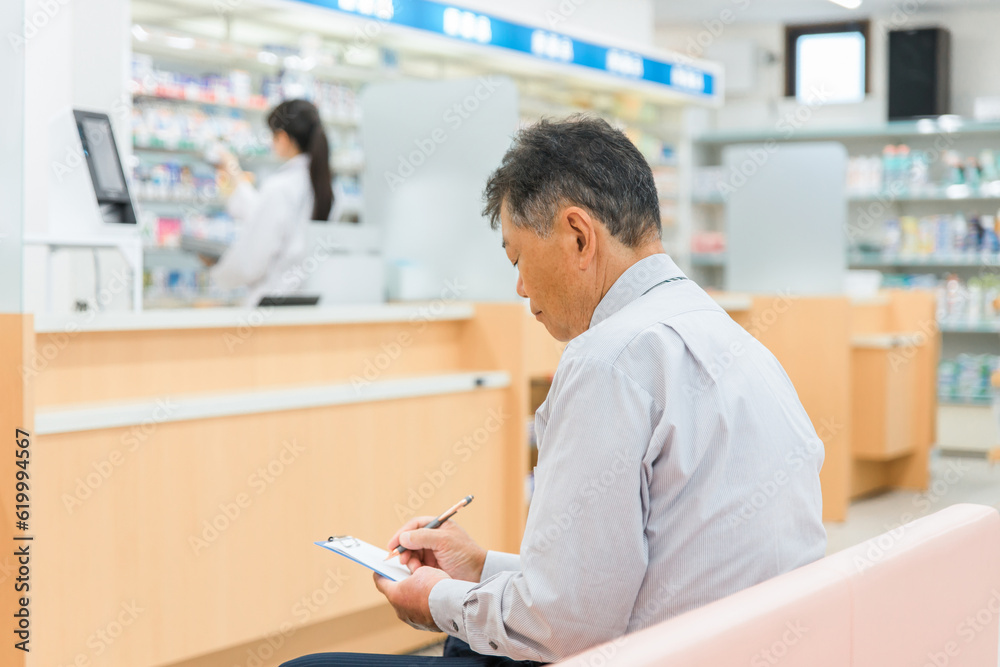 Fototapeta premium 病院・薬局の待合室で問診票を書く高齢者男性と薬剤師 