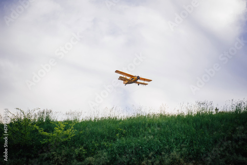 A yellow biplane flying over a populated area and a forest, spraying fertilizers for farm fields or chemicals against mosquitoes and other insects