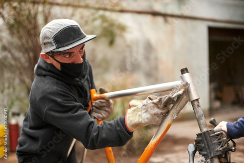 Young Hispanic man removing the paint of an orange bicycle frame, as part of a bike renovation work made at his workshop.