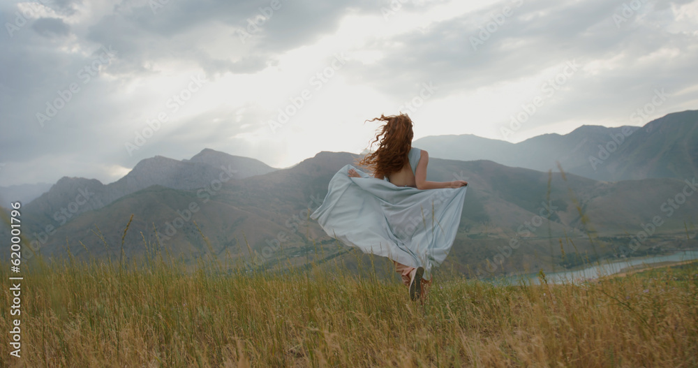 Girl in light dress with red hair is running on top of a spring mountain with scenic view. Woman in search for inspiration - freedom concept 