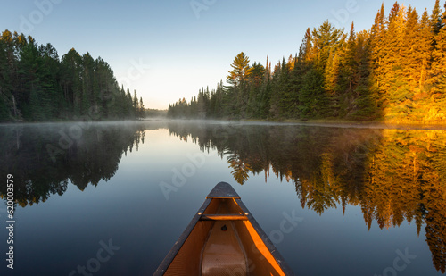 The nose of a yellow canoe sticking out in front on a glass calm late with steam and sun light hitting the tips of trees  photo