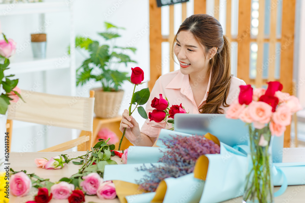 Asian professional successful female florist designer shop owner entrepreneur businesswoman sitting smiling holding decorating picking choosing red roses flowers bouquet in floral garden store studio