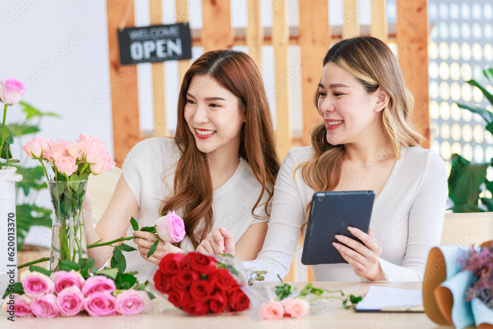 Asian professional successful female florist designer flower shop owner entrepreneur decorating pink roses bouquet in glass vase when colleague checking stock online via tablet in floral store studio