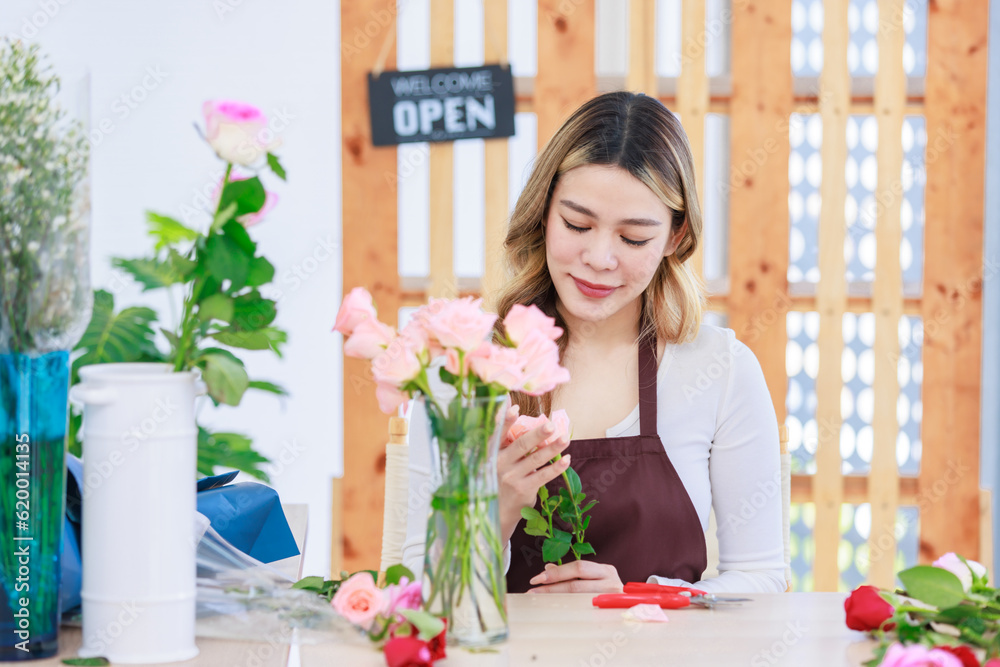 Asian happy cheerful professional female florist designer shop owner in apron sitting smiling using scissors cutting spikes decorating branch of red roses before selling in floral garden store studio Asian happy cheerful professional female florist designer shop owner in apron sitting smiling using scissors cutting spikes decorating branch of red roses before selling in floral garden store studio