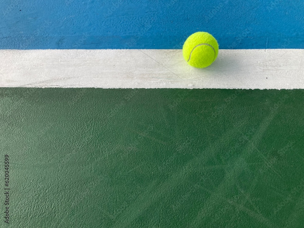 the green tennis ball is at the end of the white backline of a blue-green tennis court in daylight
