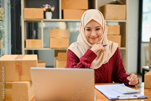 A beautiful Asian Muslim female online shop owner looking at her laptop screen