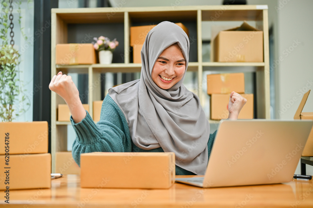 Muslim woman online business owner celebrating while looking at laptop after receiving big orders. Muslim woman online business owner celebrating while looking at laptop after receiving big orders.