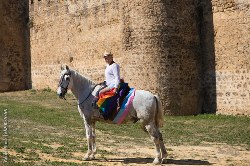Young non-binary latin person, riding a white horse with the gay pride flag. In the background a medieval castle. Concept of diversity, homosexuality and human rights.