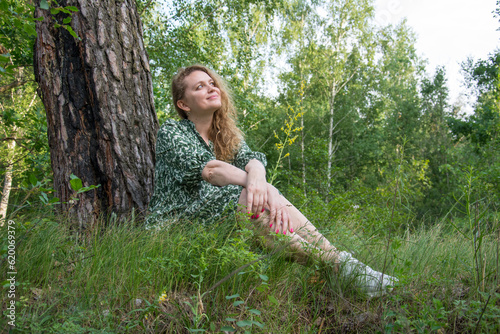 Attractive girl in the forest in a dress sits under a tree in the grass.