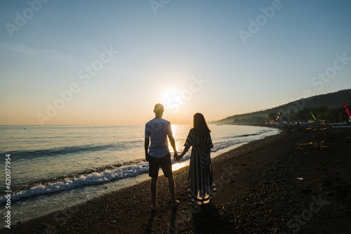 A couple of lovers, of European appearance, a man and a woman, hold hands and walk along the beach, rear view.