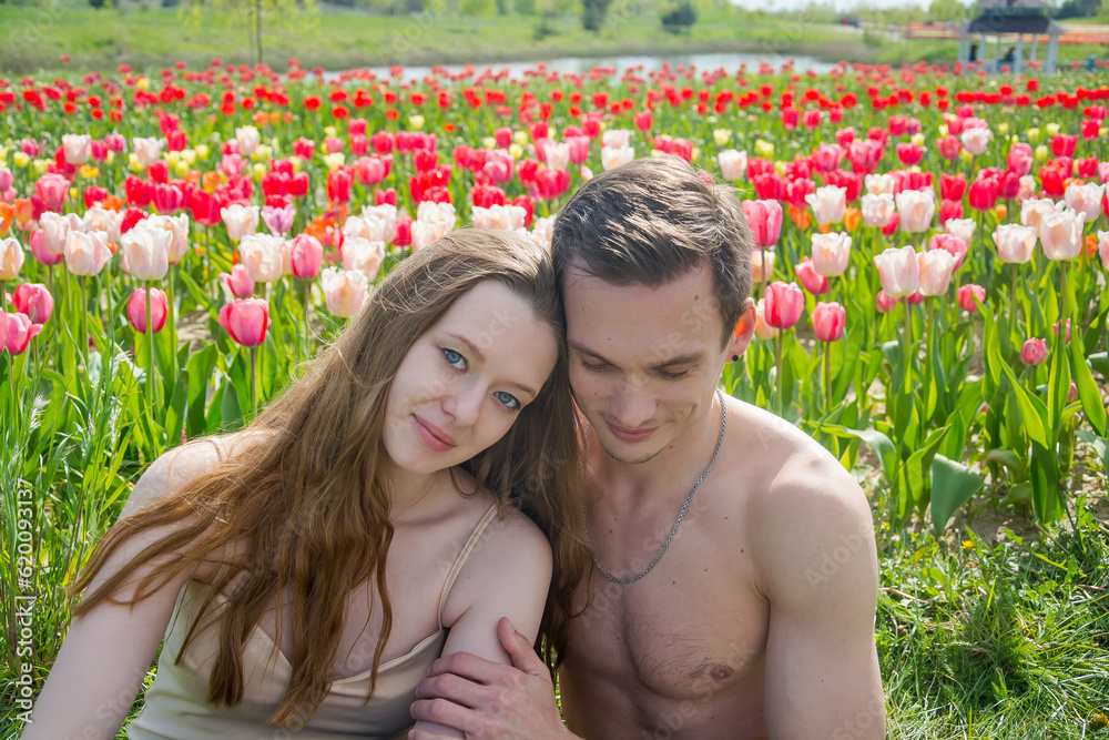 Young beautiful smiling couple sits on the background of a field of colorful tulips.