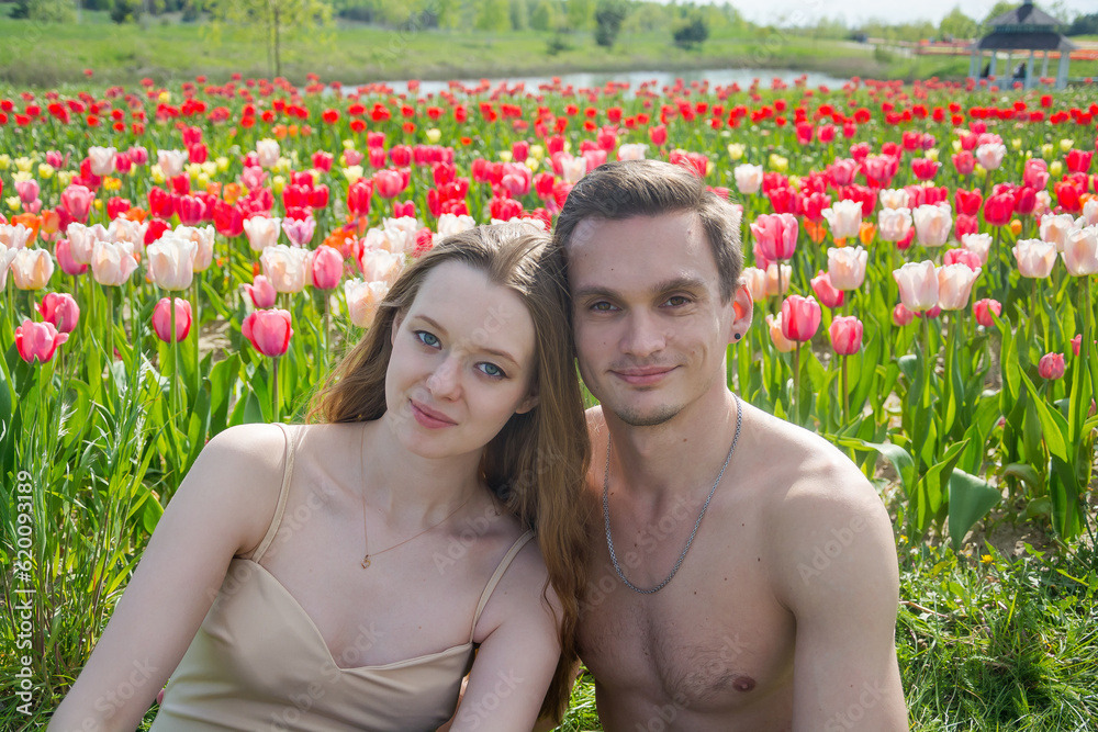 Young beautiful smiling couple sits on the background of a field of colorful tulips.