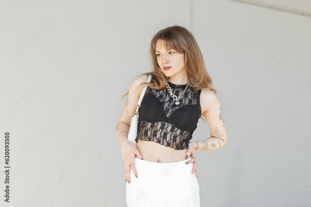 Fashionable beautiful young woman model with tattoos in a lace top with a black bra and a white skirt with a bag walks near a gray wall
