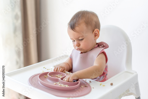Fotografie, Obraz baby little girl 8 months old sits in a high chair and eats complementary foods bulgur cereals and meat, close-up portrait looks at the camera