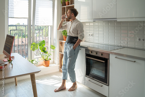Man in sick leave drinking water at home on kitchen wearing back support belt corset on lower back to treatment of hernia, relieve stress on spine, postoperative recovery. Back pain, spine problems. 