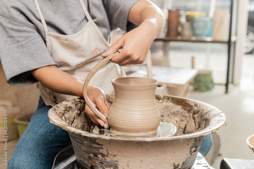 Cropped view of young female potter in apron making clay jug while working with pottery wheel in blurred ceramic workshop at background, artisanal pottery production and process Cropped view of young female potter in apron making clay jug while working with pottery wheel in blurred ceramic workshop at background, artisanal pottery production and process