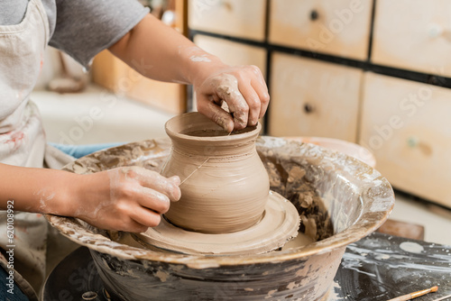 Cropped view of young female artisan in apron cutting wet clay vase on spinning pottery wheel on table in blurred ceramic workshop, clay shaping and forming process